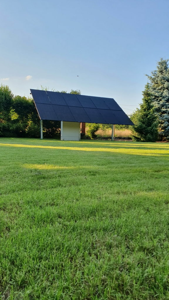 Instalacja fotowoltaiczna w formie zadaszenia nad małym budynkiem na zielonym trawniku, widoczne panele słoneczne i konstrukcja wsporcza.