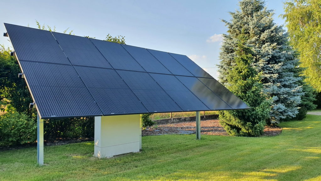 Czarny panel fotowoltaiczny na konstrukcji wsporczej, ustawiony na trawniku z widocznymi drzewami iglastymi w tle, oświetlony popołudniowym słońcem.