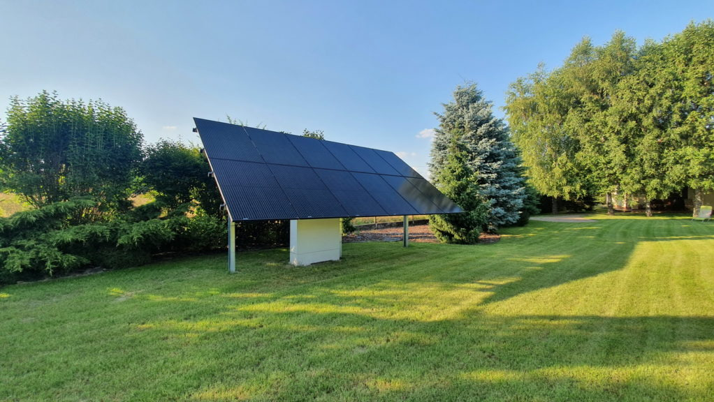 Wolnostojąca instalacja fotowoltaiczna z czarnych paneli na metalowych podporach, umieszczona na trawniku w ogrodzie, z widoczną białą skrzynką przyłączeniową i drzewami w tle.