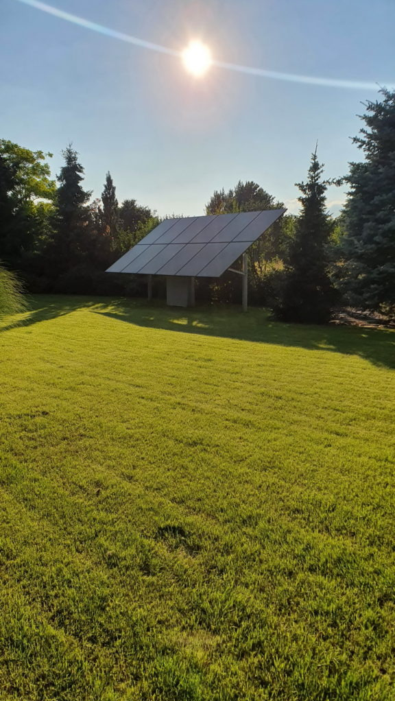 Panele słoneczne na konstrukcji wsporczej, umieszczone na trawniku z drzewami w tle, oświetlone jasnym słońcem.