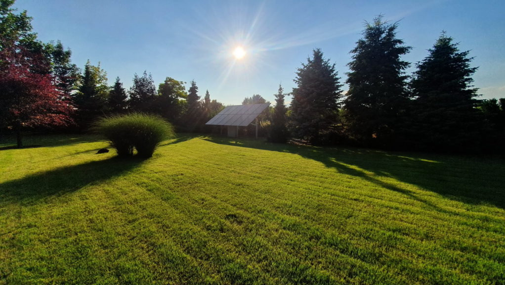 Instalacja fotowoltaiczna w ogrodzie, panele na konstrukcji wspartej na słupach, otoczone zielenią, trawnik i drzewa, słoneczny dzień.