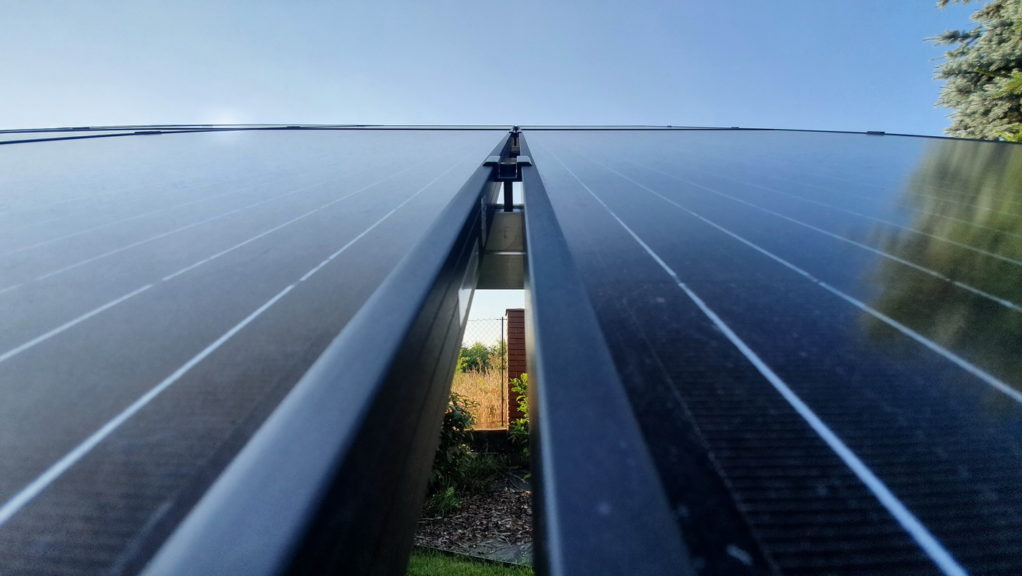 Widok z dołu na szczelinę między panelami słonecznymi, z widocznym fragmentem ogrodu i niebem w tle.