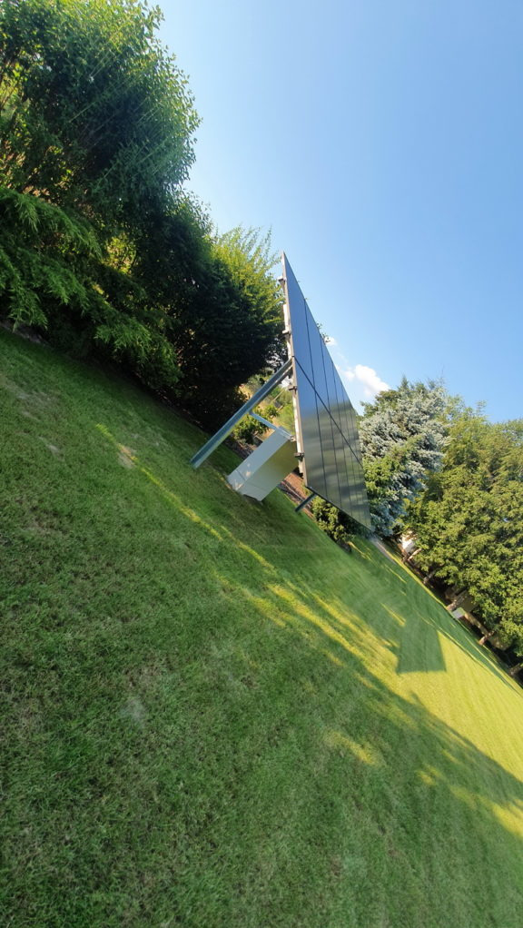 Panel słoneczny ustawiony na trawniku pod kątem, widoczne podpory i skrzynka przyłączeniowa, otoczenie zieleni drzew i krzewów, błękitne niebo w tle.