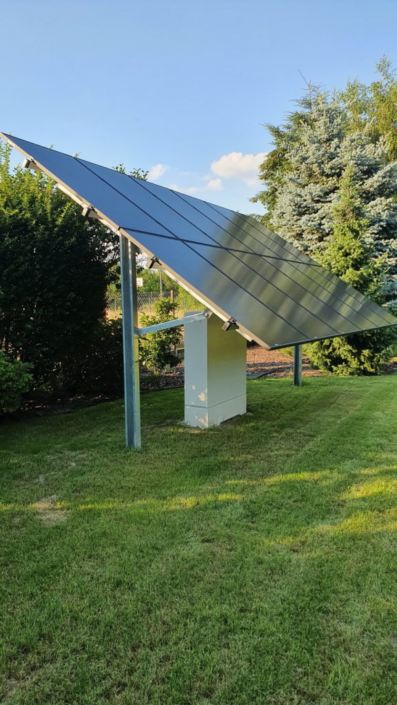 Panel fotowoltaiczny na metalowej konstrukcji, zamontowany na trawniku w ogrodzie, z białym falownikiem u podstawy, na tle drzew i błękitnego nieba.