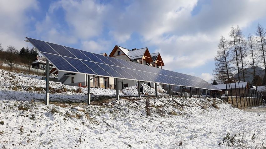 Długi rząd paneli słonecznych na metalowych podporach, zainstalowany na zasypanym śniegiem zboczu wzgórza, z budynkami w tle pod błękitnym niebem.