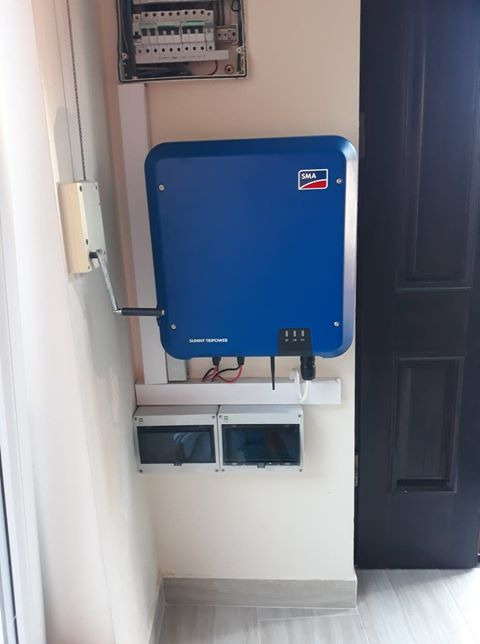 Niebieski falownik SMA Sunny Tripower zamontowany na ścianie wewnątrz budynku, z widocznymi przewodami i elementami instalacji elektrycznej, obok skrzynka bezpiecznikowa i ciemne drzwi.