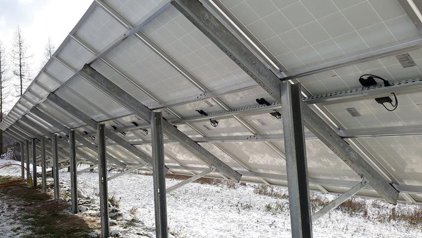 Widok od spodu na panele fotowoltaiczne zamontowane na metalowej konstrukcji, widoczne kable i łączenia, w tle ośnieżony teren.