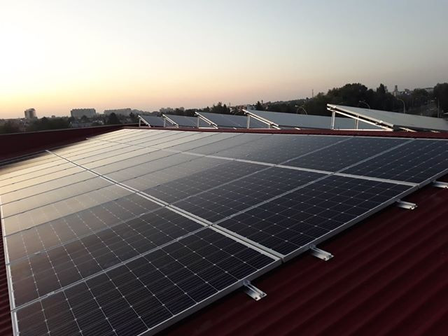 Instalacja paneli słonecznych na czerwonym, trapezowym dachu budynku, widok z lekkiego ukosa, w tle zabudowa miejska i niebo o świcie.