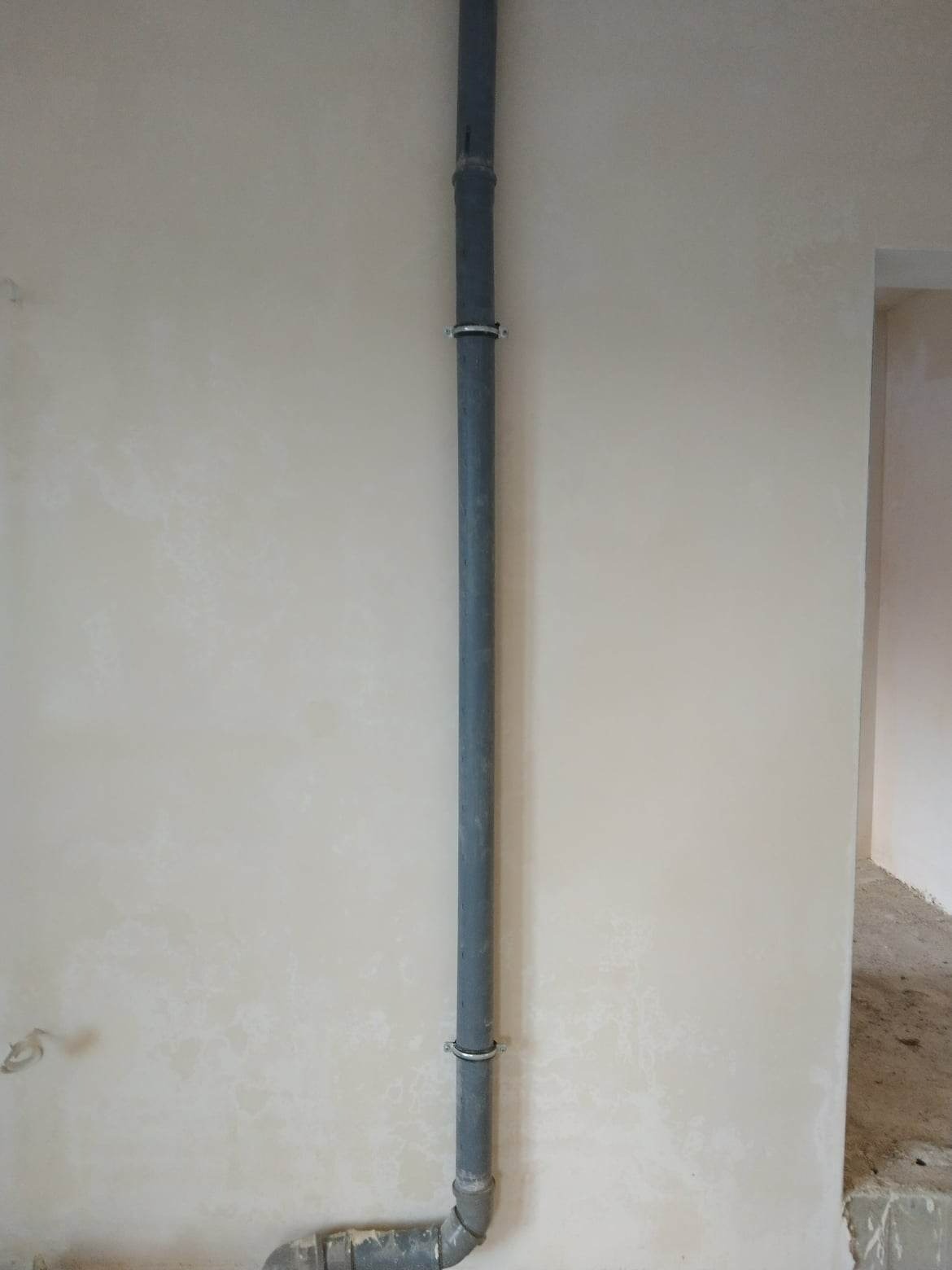 Świeżo otynkowana ściana z pionowo zamocowaną szarą rurą kanalizacyjną, widoczne metalowe obejmy mocujące, na dole kolanko łączące rurę z instalacją podłogową.
