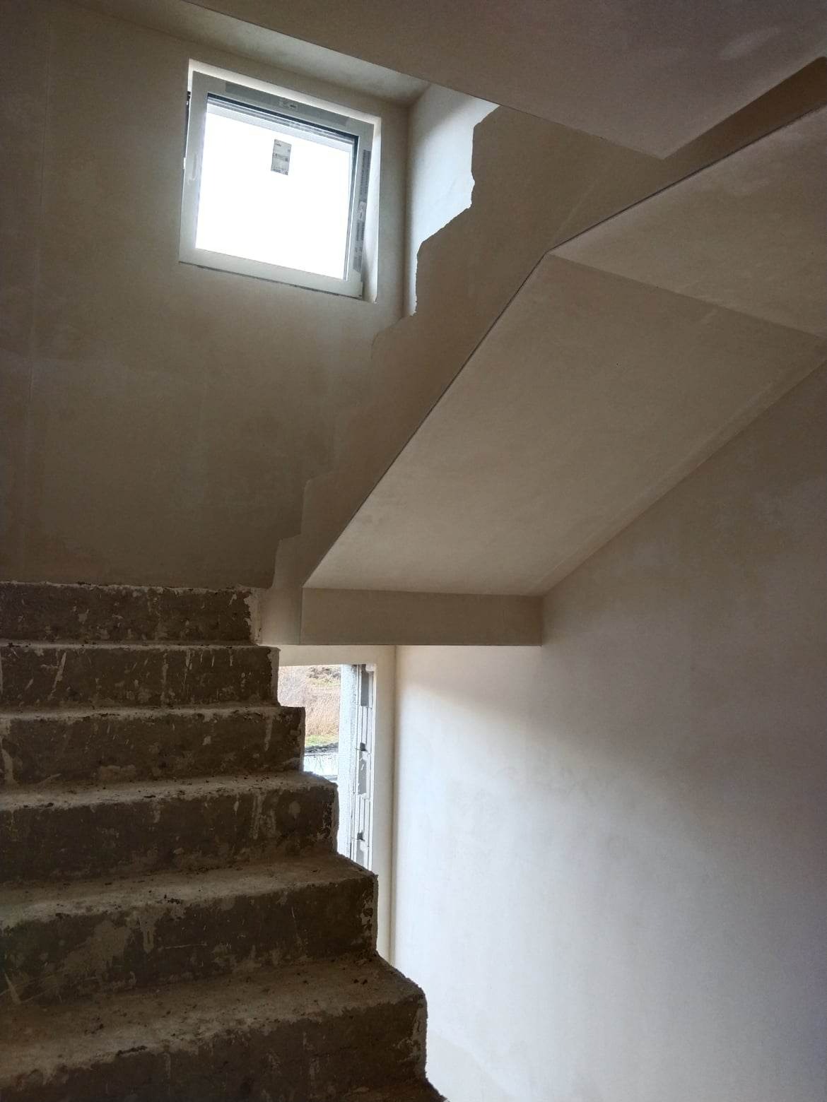 Wnętrze budynku w trakcie prac wykończeniowych: surowe schody betonowe kontrastują z gładką, jasną ścianą po wykonaniu prac tynkarskich, okno w tle