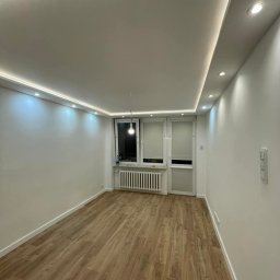 Krystian Ostaszewski - Jasne wnętrze po remoncie: panele podłogowe w odcieniach drewna, białe ściany, sufit z oświetleniem LED. Widok na okno i drzwi balkonowe. Minimalistyczny design.