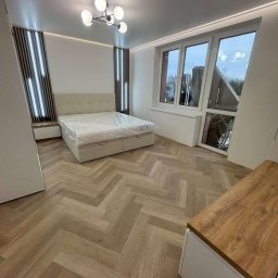 Krystian Ostaszewski - Jasna sypialnia z parkietem w jodełkę, łóżkiem z pojemnikiem i dekoracyjnymi panelami ściennymi z oświetleniem LED. Nowoczesny żyrandol i duże okno dopełniają wnętrze.