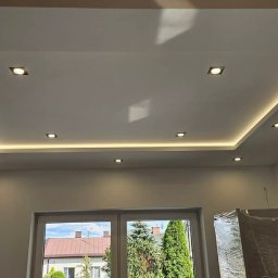 Krystian Ostaszewski - Nowoczesny sufit podwieszany z oświetleniem LED w kształcie prostokąta, z widocznym oknem i częściowo zakrytym meblem folią ochronną.