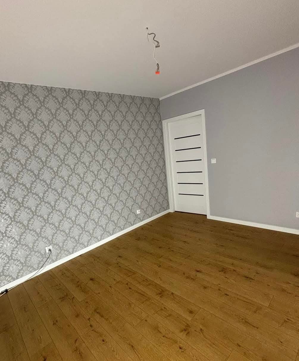 Pokój po remoncie: drewniana podłoga, ściana z wzorzystą tapetą, szara ściana, białe drzwi z poziomymi frezami. Widoczne gniazdko i wystające kable.