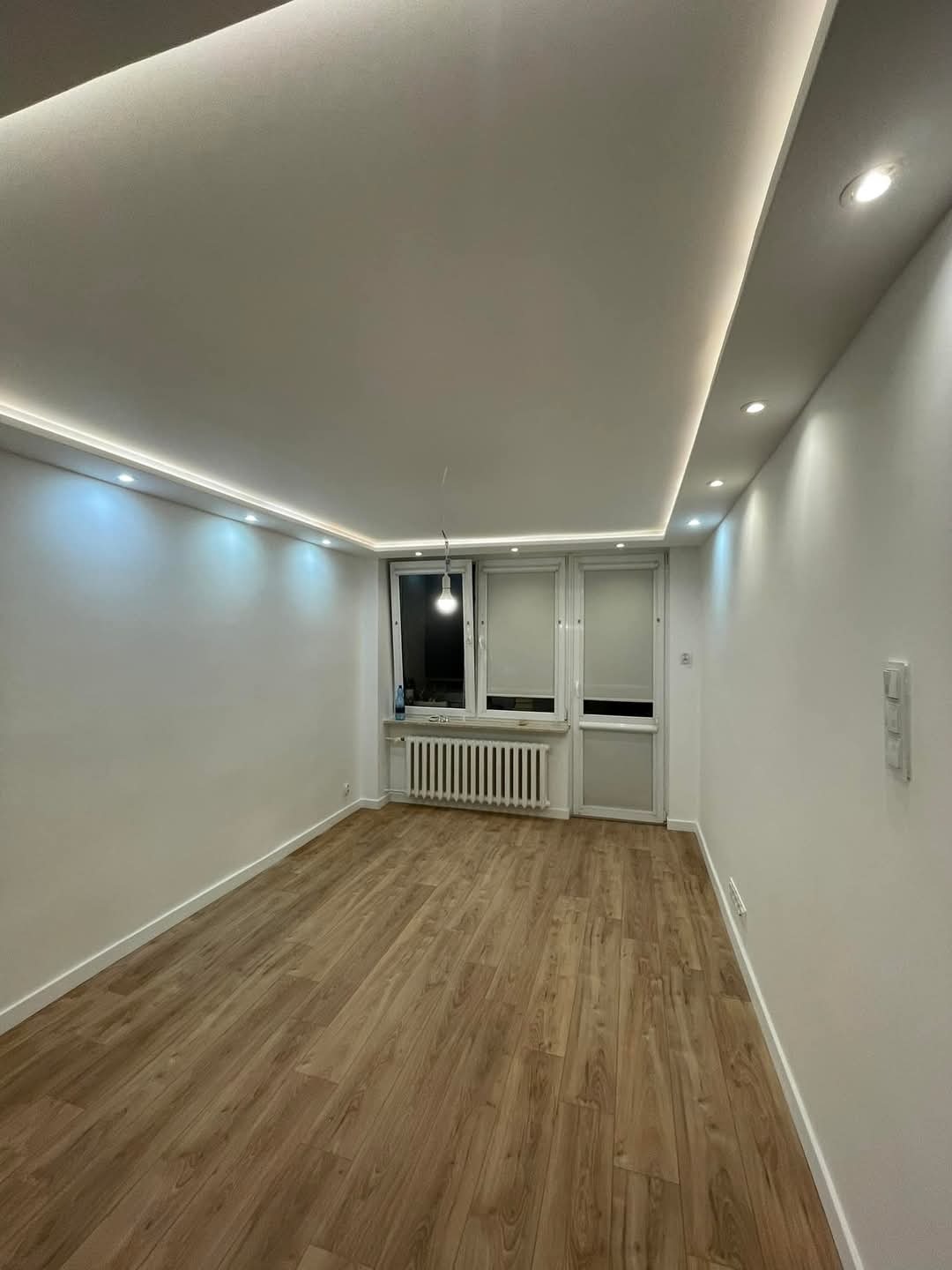 Jasne wnętrze po remoncie: panele podłogowe w odcieniach drewna, białe ściany, sufit z oświetleniem LED. Widok na okno i drzwi balkonowe. Minimalistyczny design.