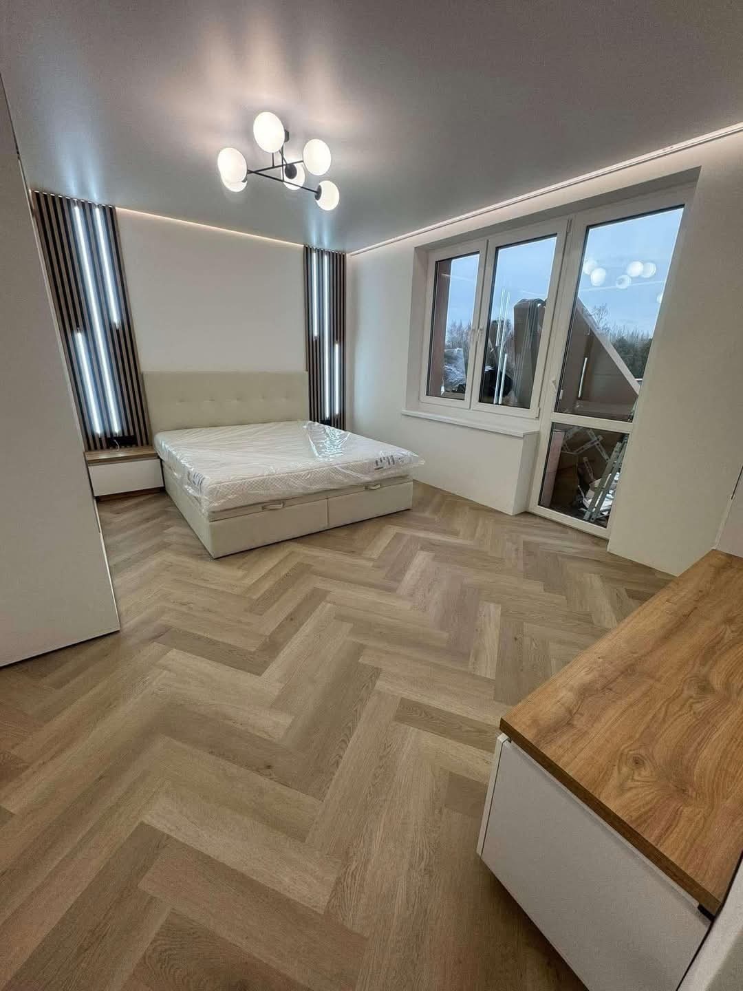 Jasna sypialnia z parkietem w jodełkę, łóżkiem z pojemnikiem i dekoracyjnymi panelami ściennymi z oświetleniem LED. Nowoczesny żyrandol i duże okno dopełniają wnętrze.