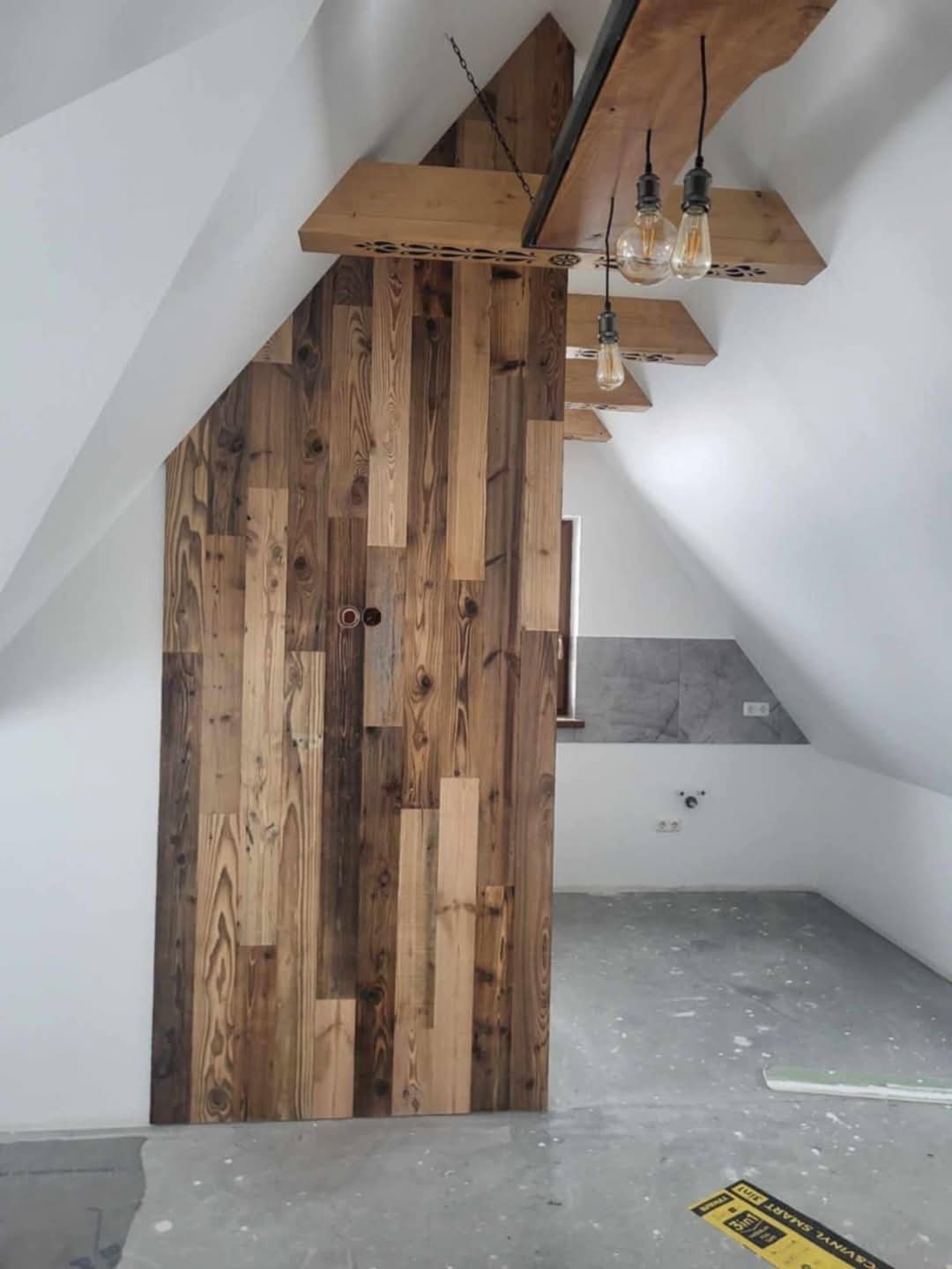 Drewniana ściana z nieregularnych desek na poddaszu, oświetlona wiszącymi lampami. Widoczny fragment łazienki z szarą glazurą. Białe ściany i sufit.