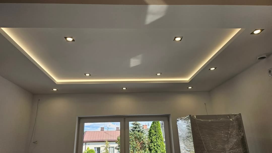 Nowoczesny sufit podwieszany z oświetleniem LED w kształcie prostokąta, z widocznym oknem i częściowo zakrytym meblem folią ochronną.