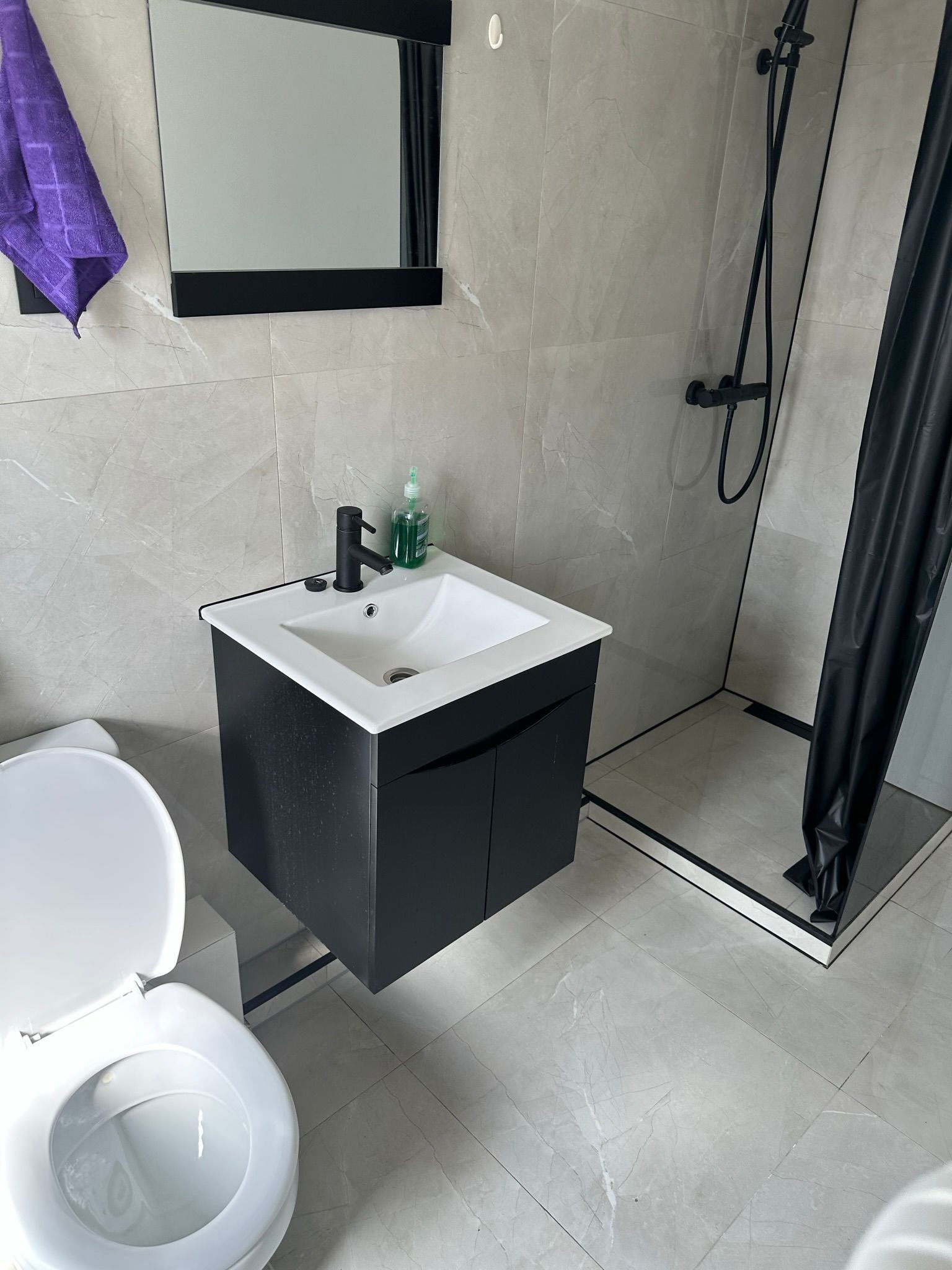 Nowoczesna łazienka z szarymi płytkami, czarną armaturą i szafką pod umywalką. Widoczny fragment toalety i kabina prysznicowa z czarną zasłoną. Minimalistyczny design.