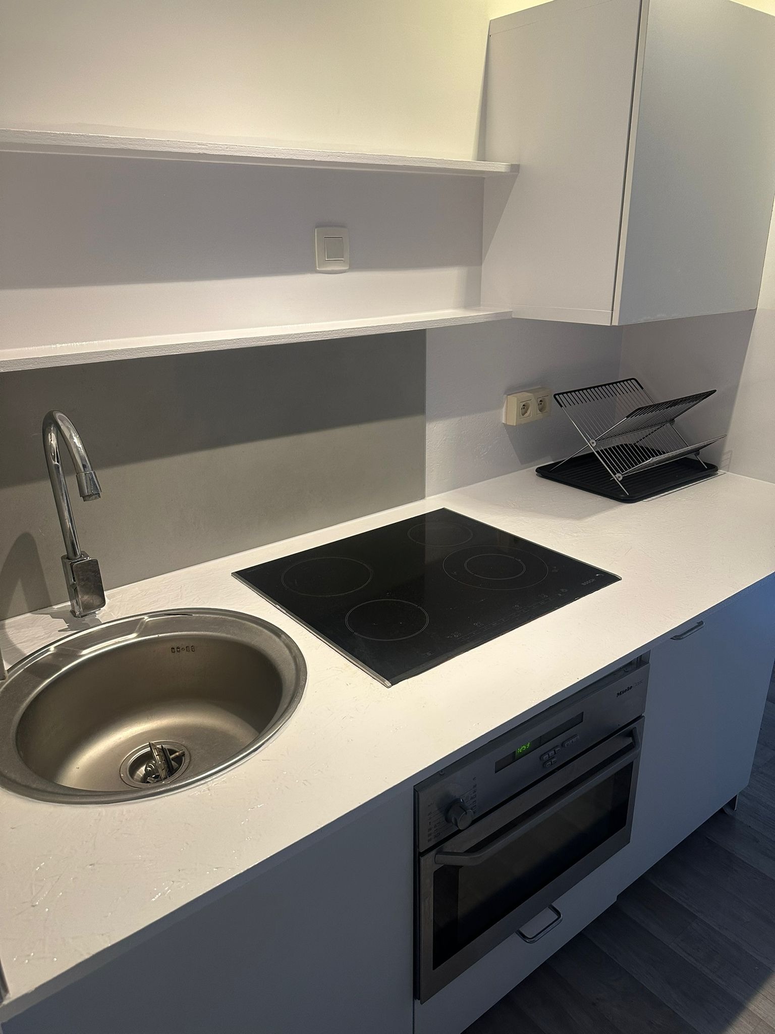 Nowoczesna, minimalistyczna kuchnia w odcieniach bieli. Widoczny zlew, płyta indukcyjna, piekarnik Miele i suszarka do naczyń. Proste półki i szafki.