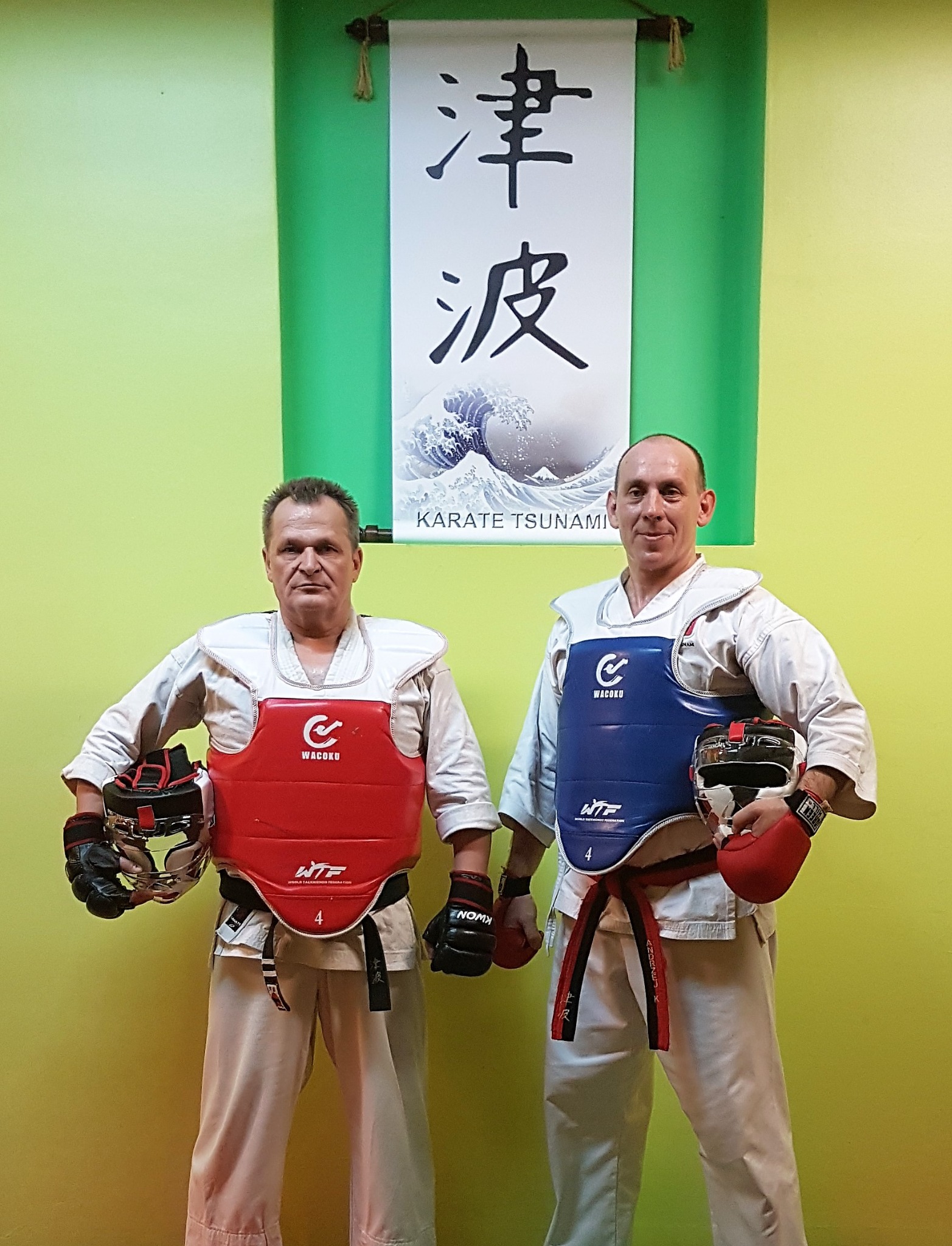 Dwóch mężczyzn w strojach do sportów walki, z ochraniaczami na korpus (czerwony i niebieski) i rękawicami, pozuje na tle zielonej ściany z japońskim zwojem 'Karate Tsunami'.