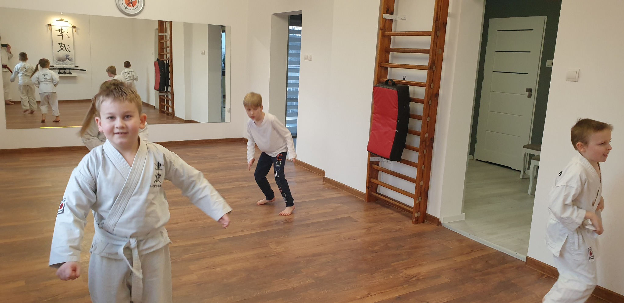 Chłopiec w białym kimonie karate pozuje na tle sali treningowej z drewnianą podłogą, drabinkami i lustrem odbijającym grupę dzieci podczas zajęć.