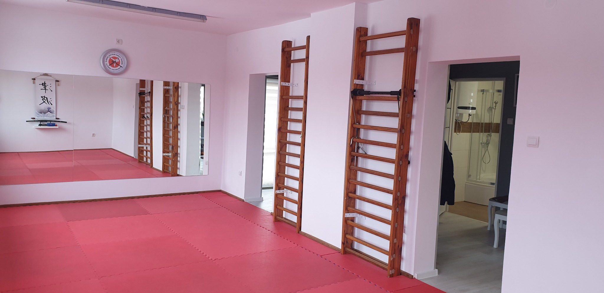Sala do ćwiczeń z czerwonymi matami na podłodze, lustrem na ścianie i drabinkami gimnastycznymi; widok na łazienkę w tle.