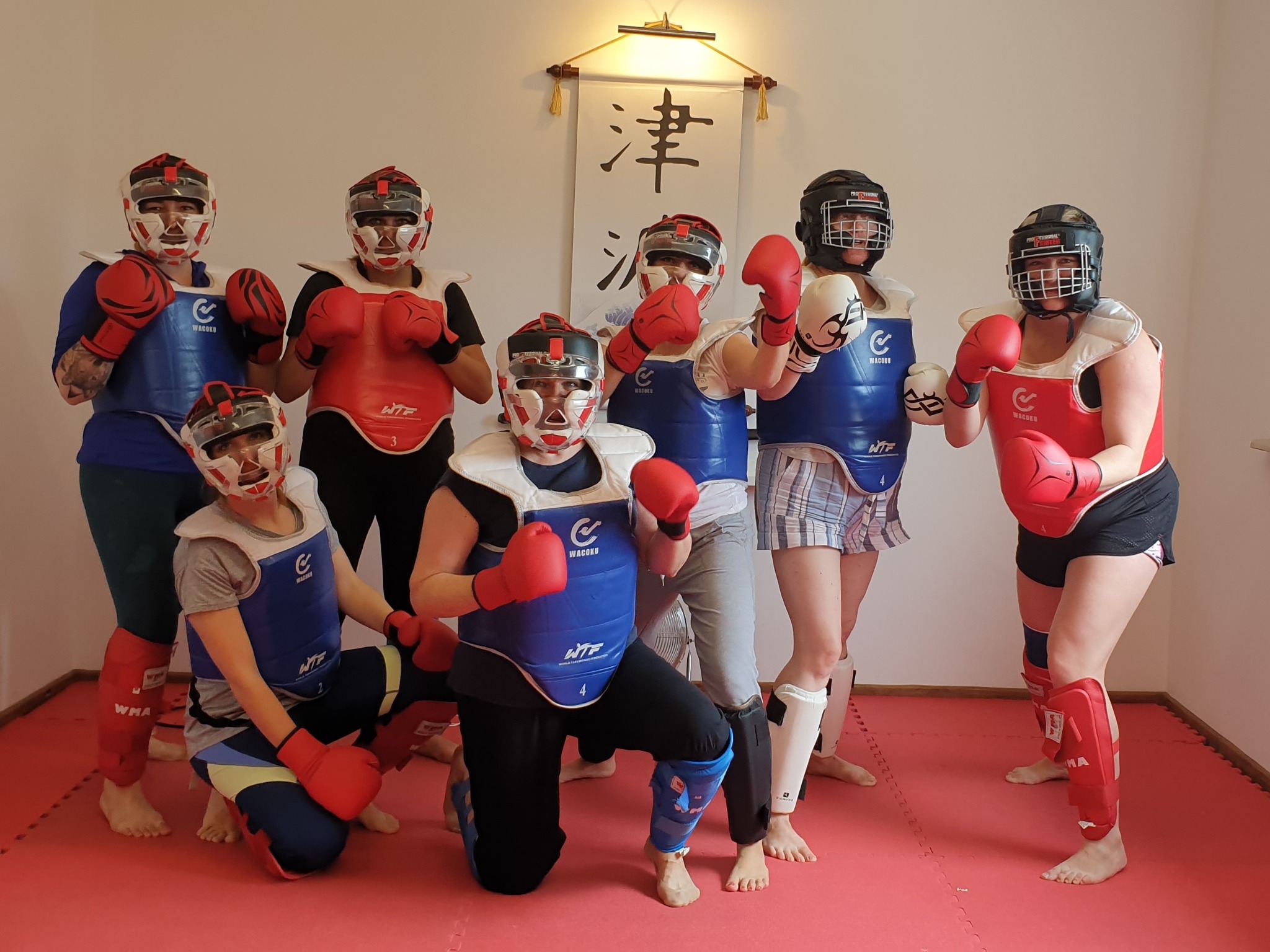 Grupa ośmiu osób w strojach ochronnych do sportów walki, w tym kaski, ochraniacze na korpus i rękawice, pozuje na czerwonej macie w pomieszczeniu z kaligrafią na ścianie.