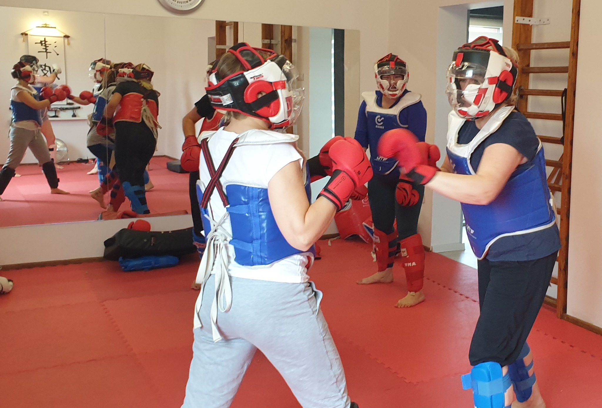 Grupa osób w kaskach i ochraniaczach, ćwiczących sporty walki na sali z czerwonym parkietem, widoczne odbicie w lustrze.