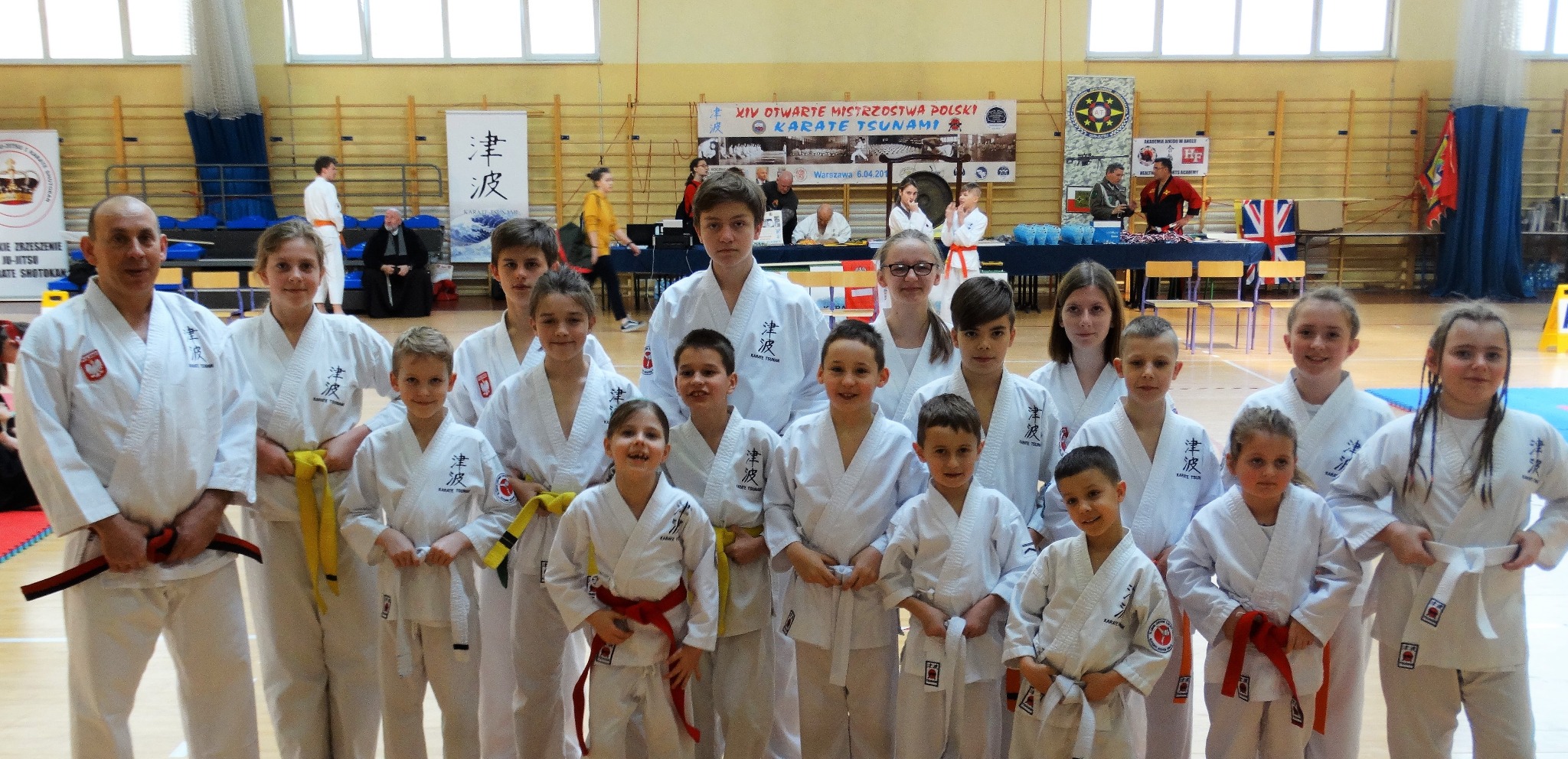 Grupa dzieci i dorosły w białych kimonach z różnymi kolorami pasów, stojąca na tle sali gimnastycznej podczas zawodów karate, widoczny baner z napisem XIV Otwarte Mistrzostwa Polski Karate Tsunami.