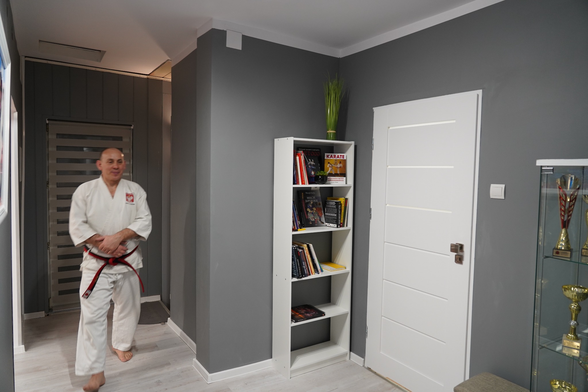 Mężczyzna w kimonie z czerwonym pasem, zbliżający się w kierunku kamery, na tle wnętrza z regałem pełnym książek o tematyce karate i sportów walki, widoczne trofea w szklanej gablocie.
