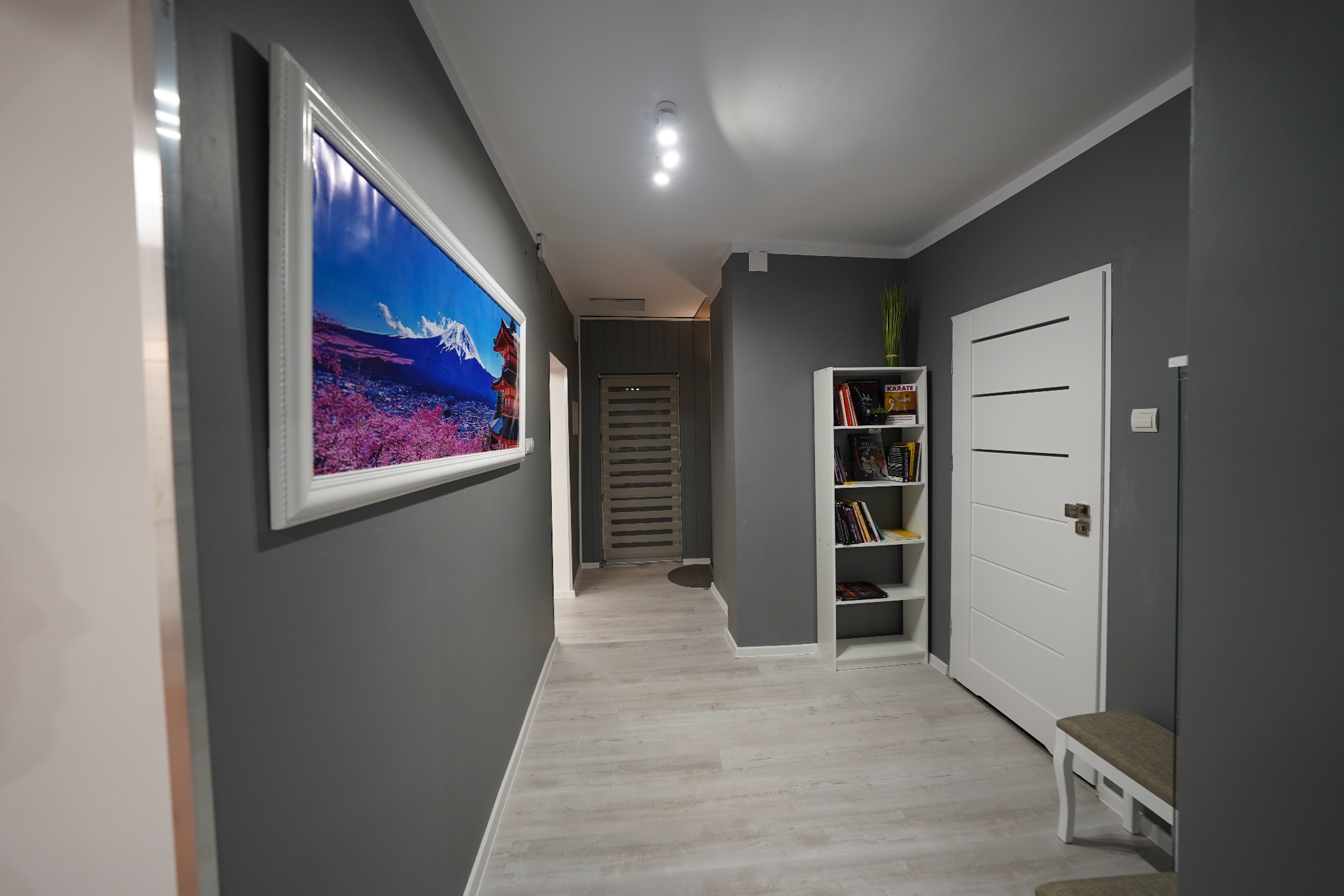 Nowoczesny korytarz z szarą ścianą, na której wisi obraz z motywem góry Fuji w ozdobnej ramie, białe drzwi z poziomymi frezowaniami i biały regał z książkami.