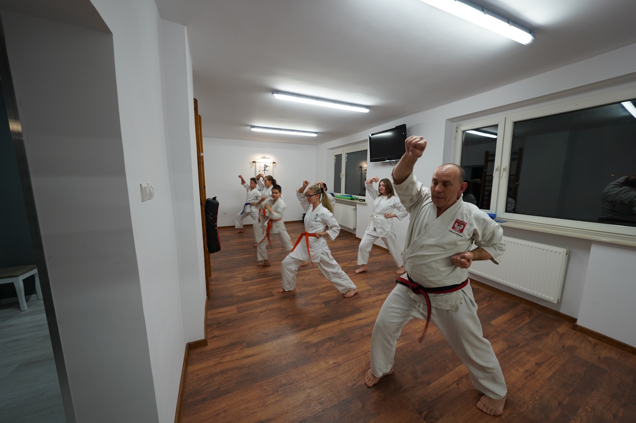 Sala do sztuk walki z grupą osób w kimonach, wykonujących ćwiczenia. Instruktor w białym kimonie z czarnym pasem na pierwszym planie, unosi pięść do góry. Na kimonie widoczna naszywka z polską...