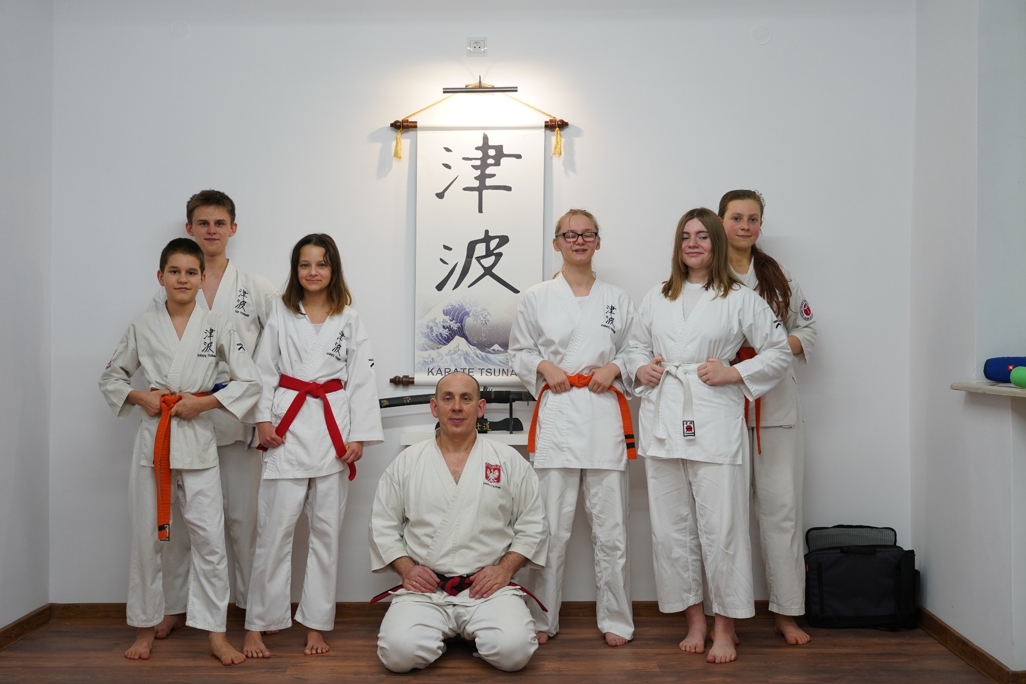 Grupa uczniów karate w białych kimonach z kolorowymi pasami, pozujących z instruktorem na tle ściany z japońskim zwojem.