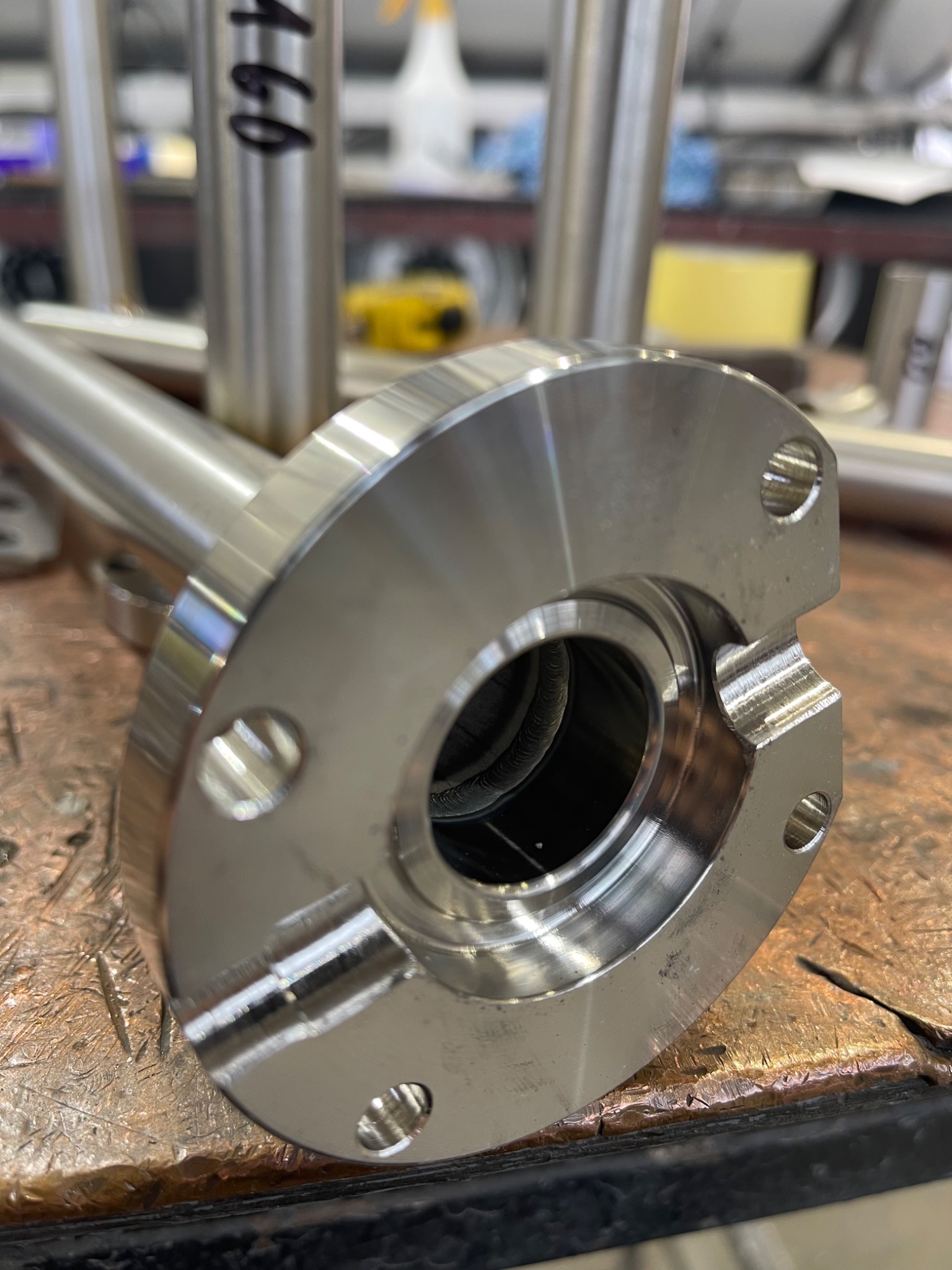 Wypolerowany, metalowy element z kołnierzem i widocznym spawem wewnątrz otworu, leżący na metalowej powierzchni roboczej.