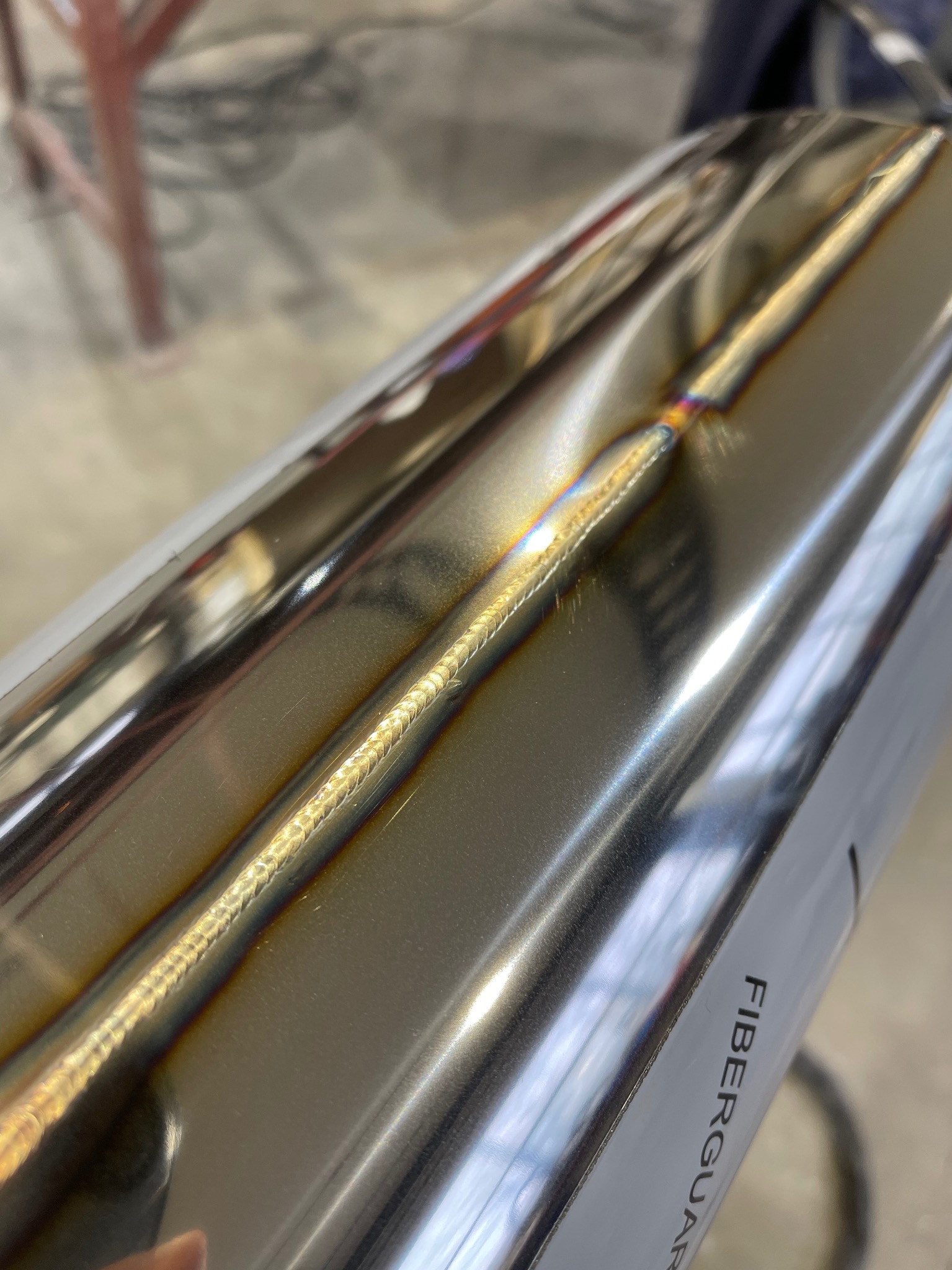 Błyszcząca, spawana rura o przekroju kwadratowym, z widocznym, precyzyjnym szwem spawalniczym i opalizującymi przebarwieniami termicznymi, z napisem 'FIBERGUAR' wzdłuż jednej krawędzi.