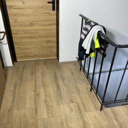 ALLIN1 - Beżowe panele podłogowe imitujące drewno, brązowe drzwi z czarną klamką, czarna balustrada schodów z powieszoną kurtką.