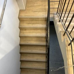 ALLIN1 - Widok z góry na schody wykończone panelami drewnopodobnymi, z czarną metalową balustradą po prawej stronie i białymi ścianami po lewej, fragment sufitu w tle.