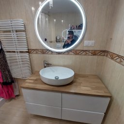 ALLIN1 - Nowoczesna łazienka z okrągłym lustrem podświetlanym LED, białą umywalką nablatową na drewnianym blacie i białymi szafkami pod umywalką. W tle widoczny grzejnik drabinkowy i pralka.