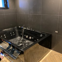 ALLIN1 - Czarna wanna z hydromasażem, z chromowanymi elementami sterującymi i dyszami, zamontowana w łazience z ciemnymi, kwadratowymi płytkami, częściowo widoczna podłoga z jasnego drewna ułożonego w jodełkę.