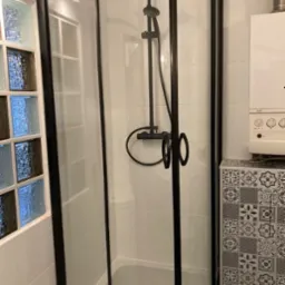 Kabina prysznicowa narożna z czarnymi profilami, białe płytki na ścianach, dekoracyjne luksfery i wzorzysta zabudowa obok kotła.