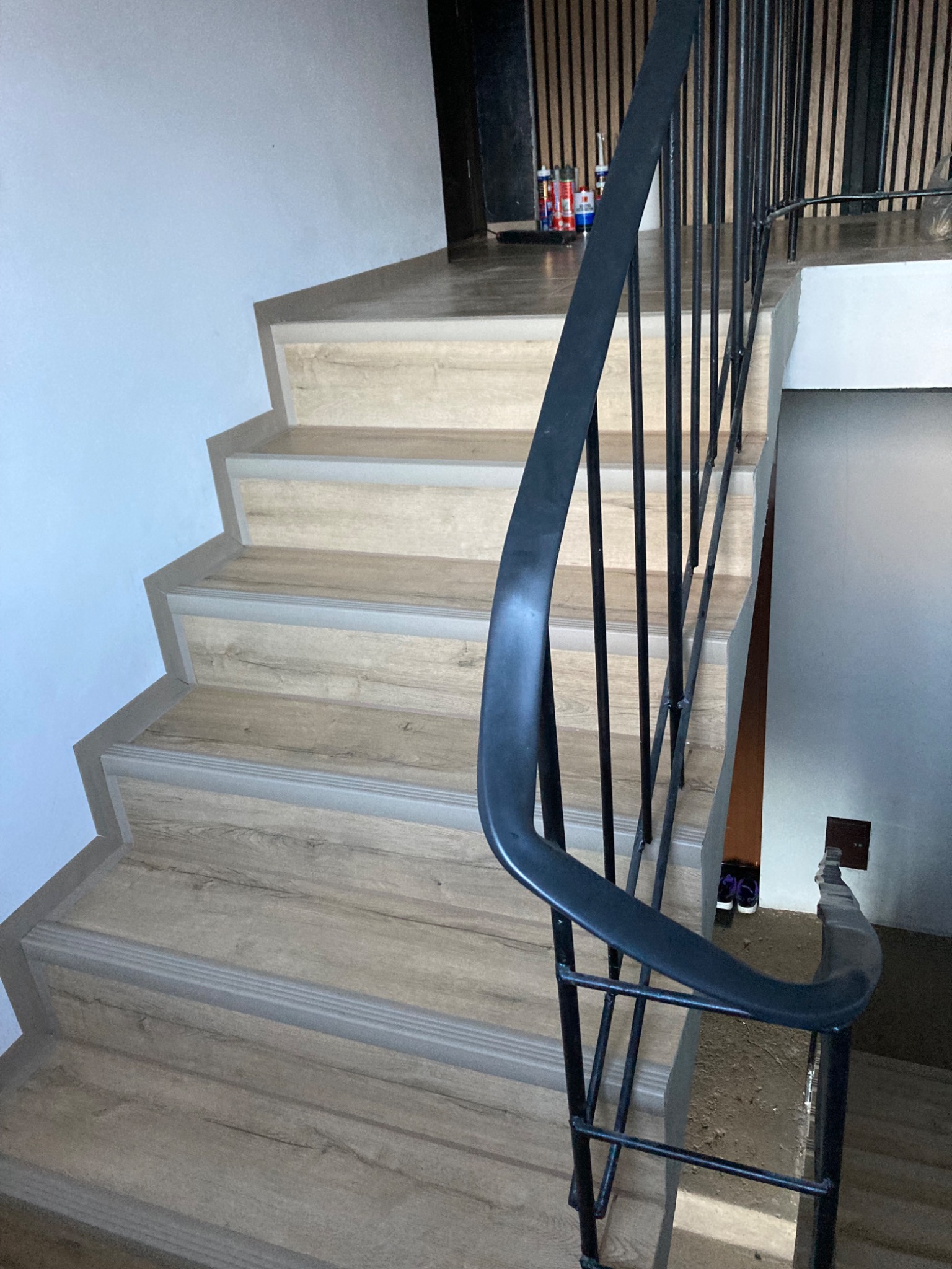 Schody wykończone panelami drewnopodobnymi z szarą listwą przy krawędzi, czarna metalowa balustrada, widoczne farby i silikony w tle.