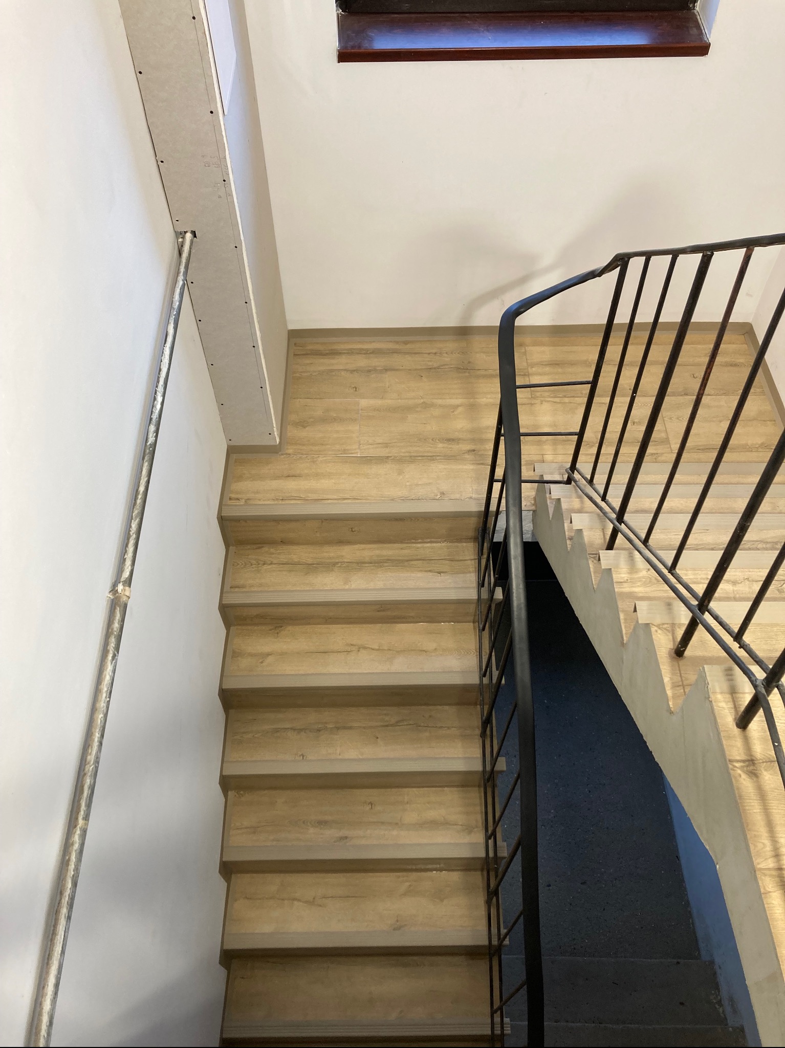 Widok z góry na klatkę schodową z drewnopodobnymi panelami na stopniach, czarną metalową balustradą i fragmentem ściany z zamontowaną płytą kartonowo-gipsową oraz metalowym profilem.