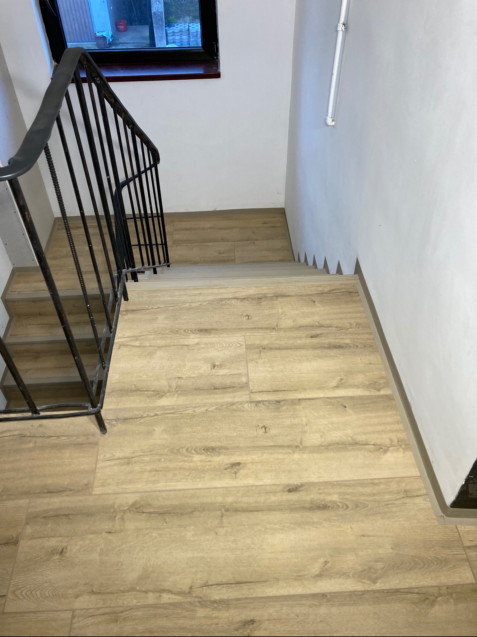 Schody wyłożone panelami podłogowymi o jasnym odcieniu drewna, widok z góry, z czarną metalową barierką po lewej stronie i białymi ścianami.