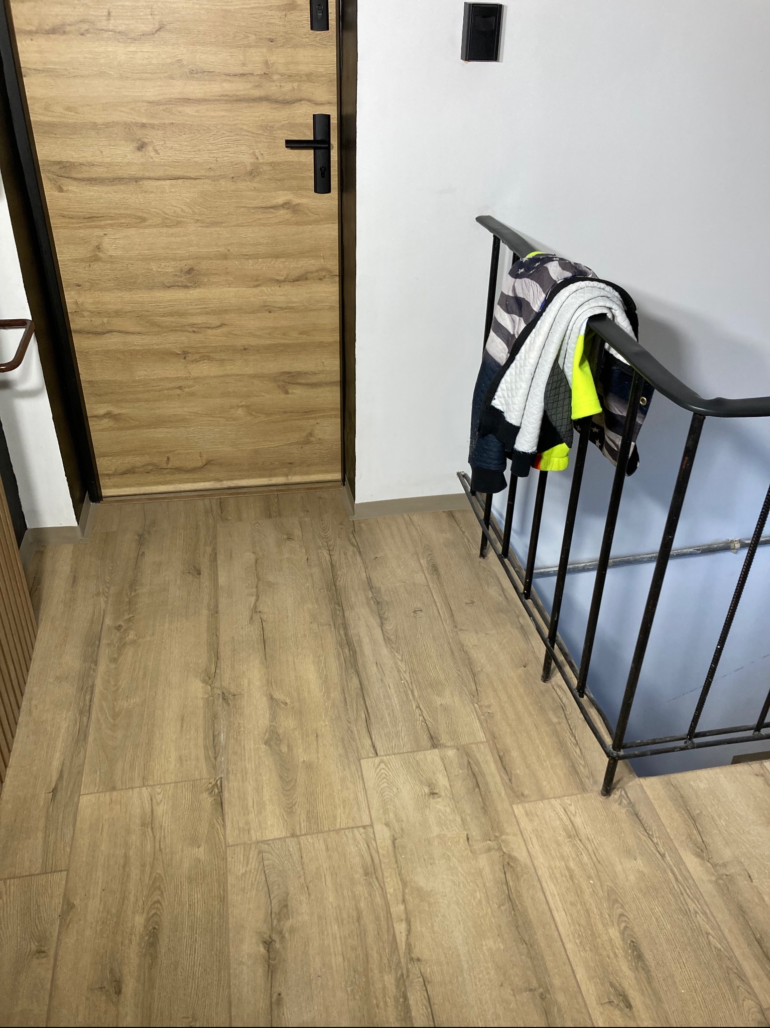 Beżowe panele podłogowe imitujące drewno, brązowe drzwi z czarną klamką, czarna balustrada schodów z powieszoną kurtką.