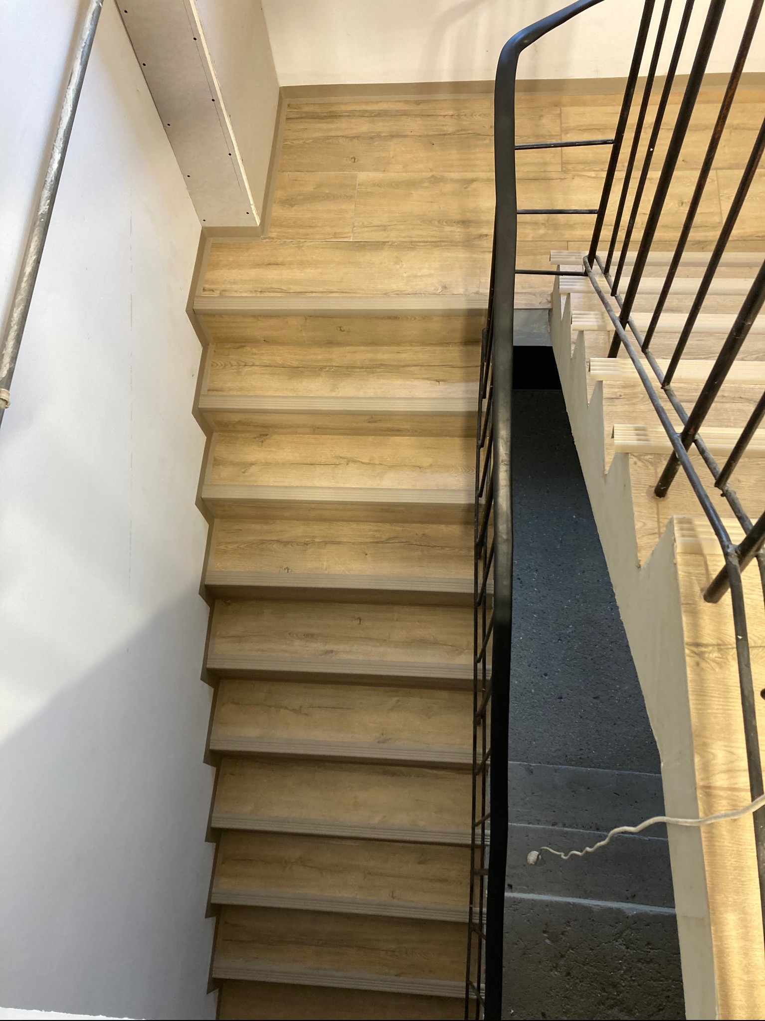 Widok z góry na schody wykończone panelami drewnopodobnymi, z czarną metalową balustradą po prawej stronie i białymi ścianami po lewej, fragment sufitu w tle.