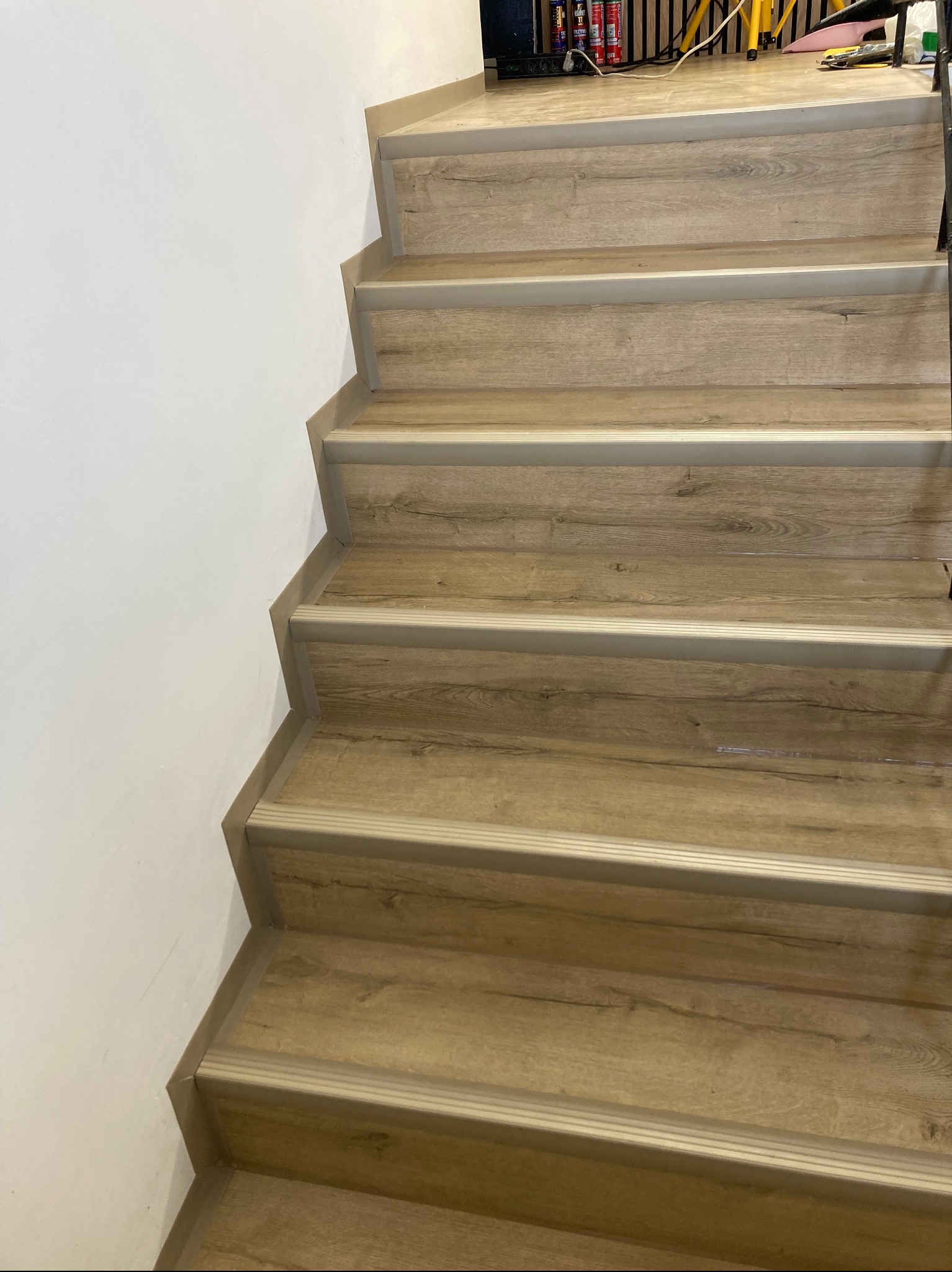 Schody wykończone panelami imitującymi jasne drewno, zabezpieczone srebrnymi listwami krawędziowymi, przylegające do białej ściany.