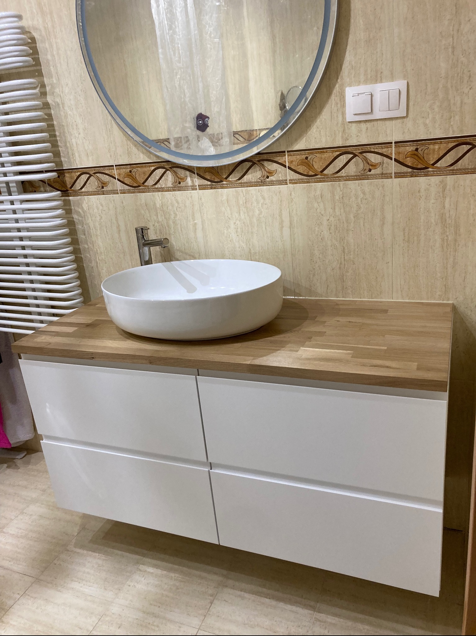 Umywalka nablatowa w kształcie misy na szafce z drewnianym blatem w łazience z beżowymi płytkami i okrągłym lustrem z podświetleniem LED.