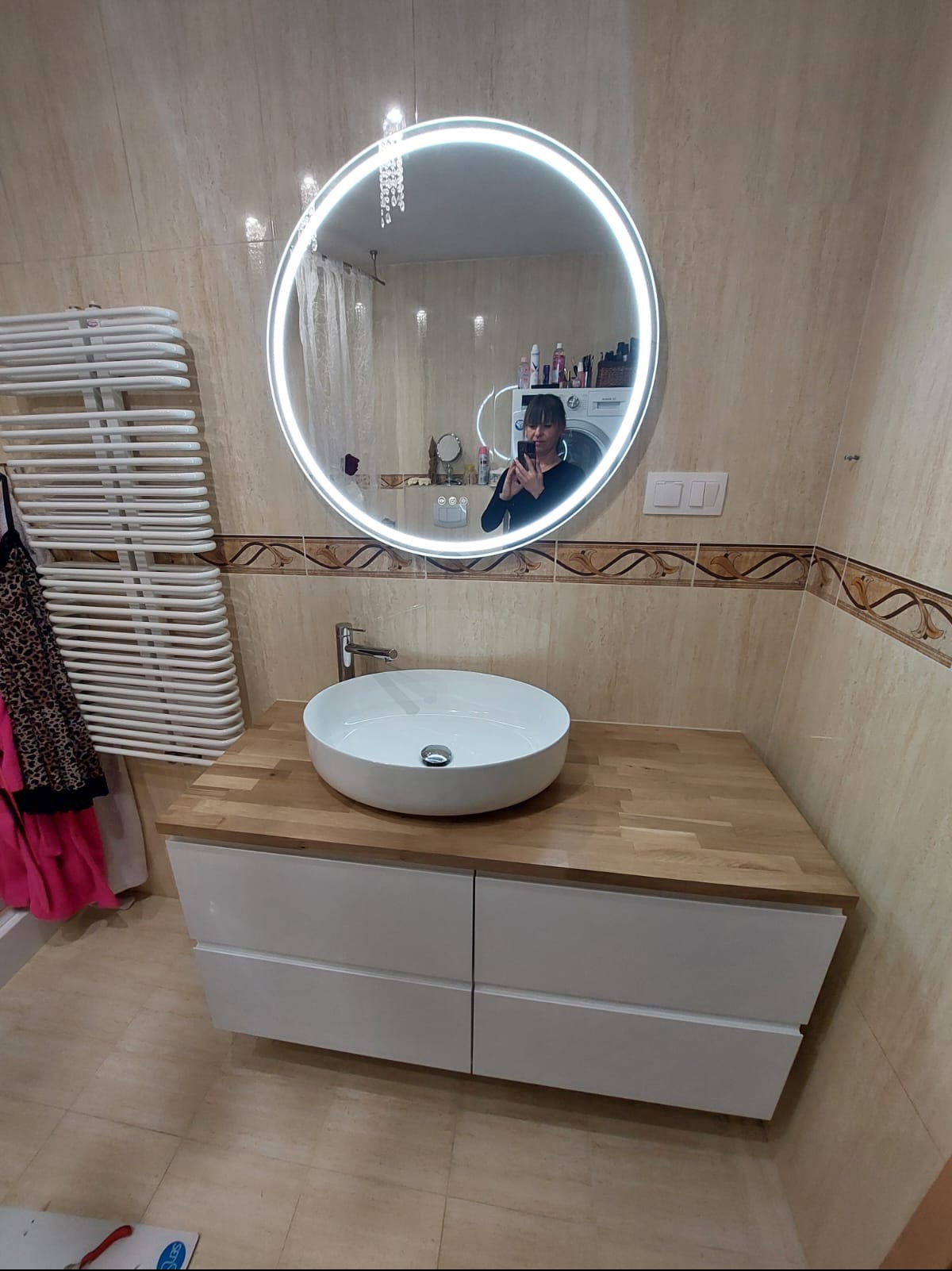 Nowoczesna łazienka z okrągłym lustrem podświetlanym LED, białą umywalką nablatową na drewnianym blacie i białymi szafkami pod umywalką. W tle widoczny grzejnik drabinkowy i pralka.