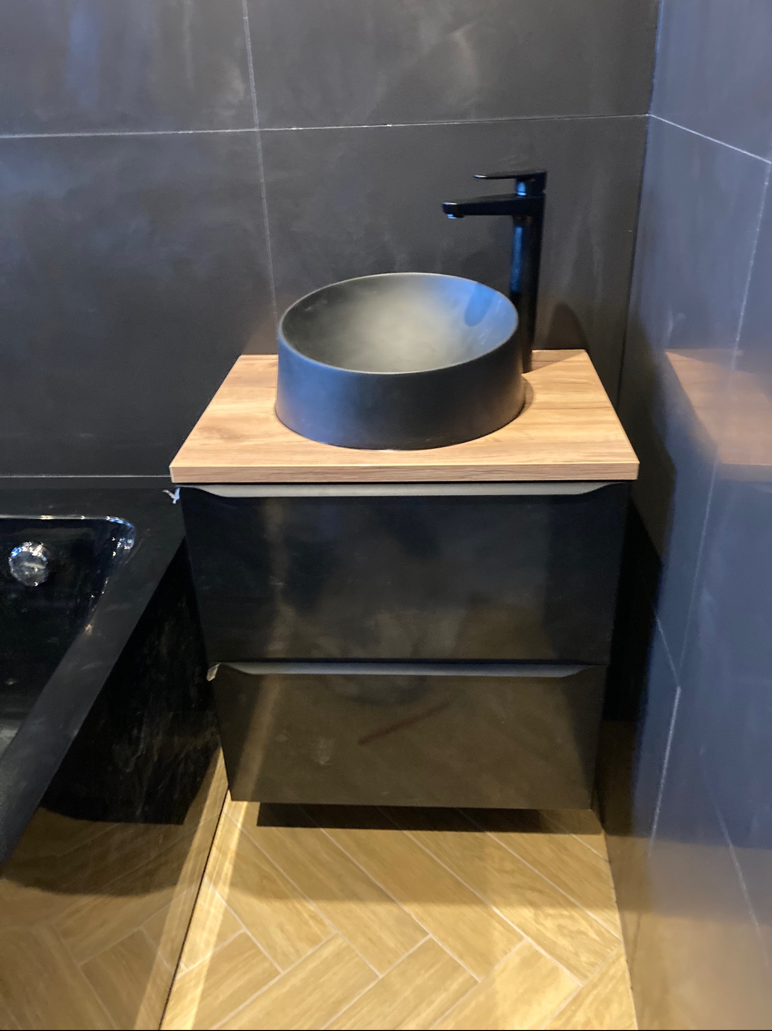 Nowoczesna łazienka z czarną umywalką nablatową, drewnianym blatem i ciemnymi płytkami na ścianach, uzupełniona jasną podłogą w jodełkę.